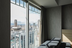 Habitación de hotel moderna con un ventanal grande que ofrece vistas a la ciudad, cabecera de madera y un banco integrado.