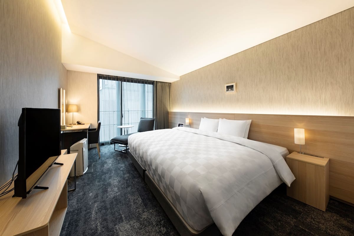 A hotel room with a large bed, a desk with a TV, a chair, and a small table.