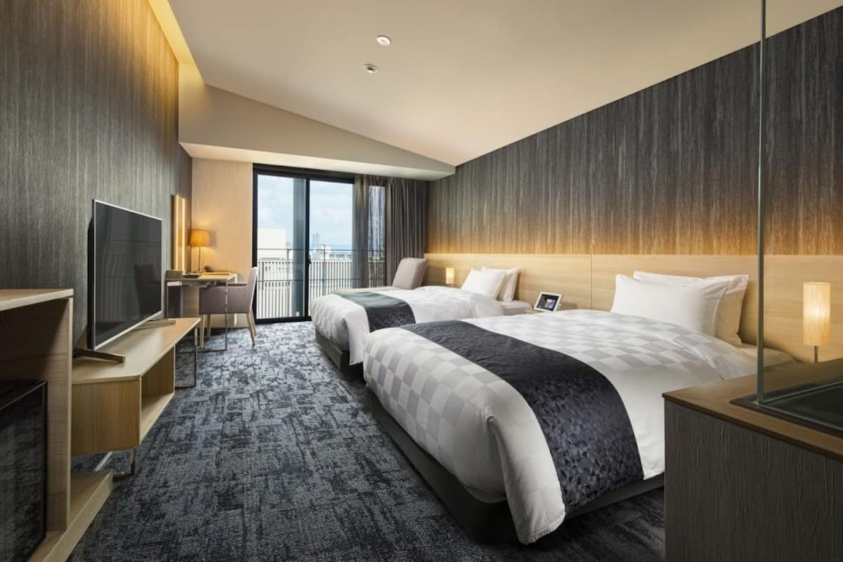 A modern hotel room with two beds, a desk, a TV, and a balcony with a city view.
