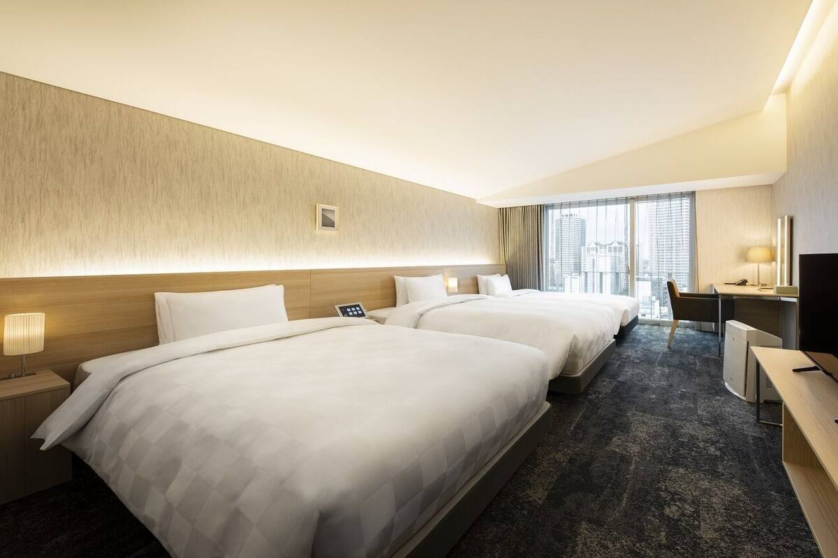 A hotel room with two beds, a desk, a TV, and a city view.