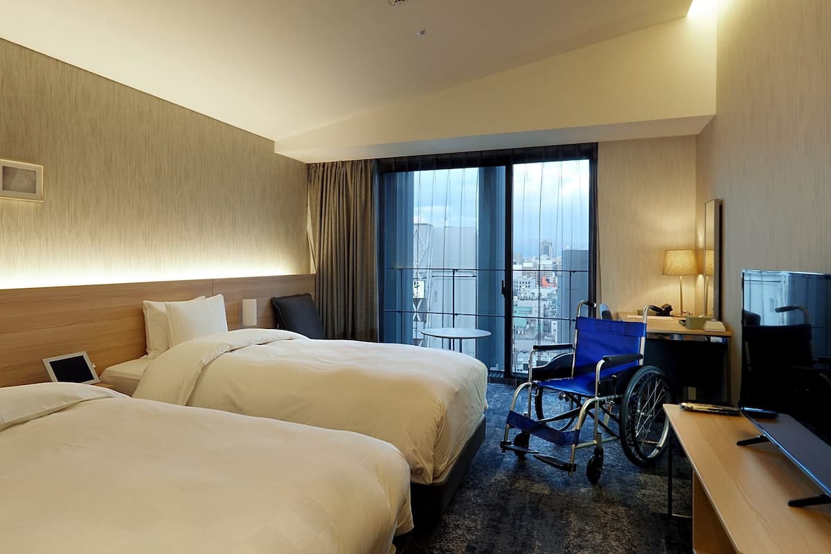 A hotel room with two beds, a desk with a chair, a wheelchair, and a large window with a view of buildings.