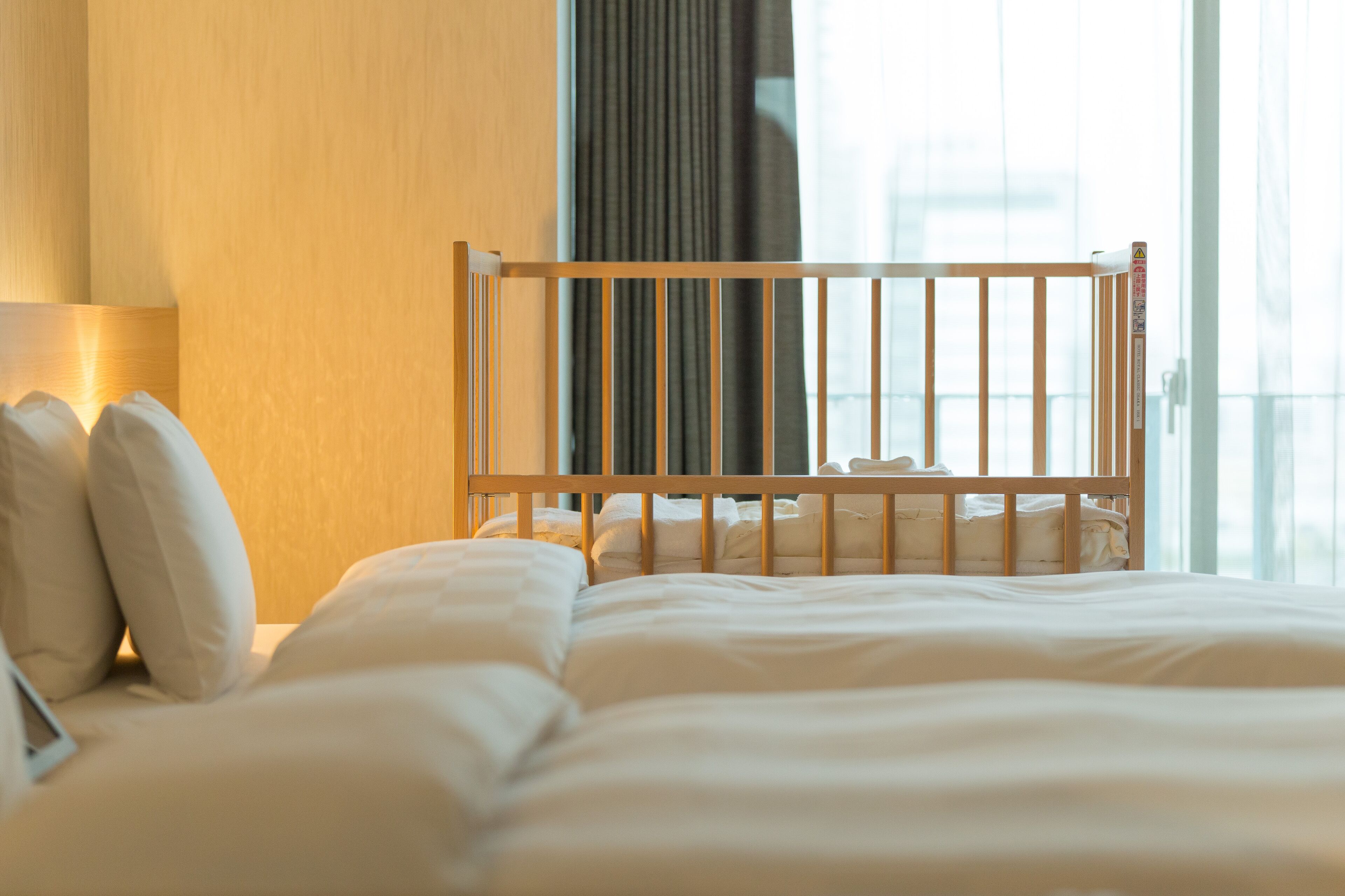 A hotel room with a wooden crib, a bed with white bedding, a bedside lamp, and a view of the city through large windows.