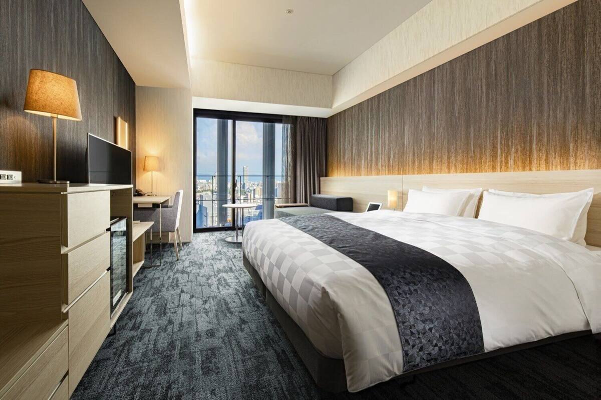 A modern hotel room with a large bed, a desk, a chair, and a view of the city through large windows.