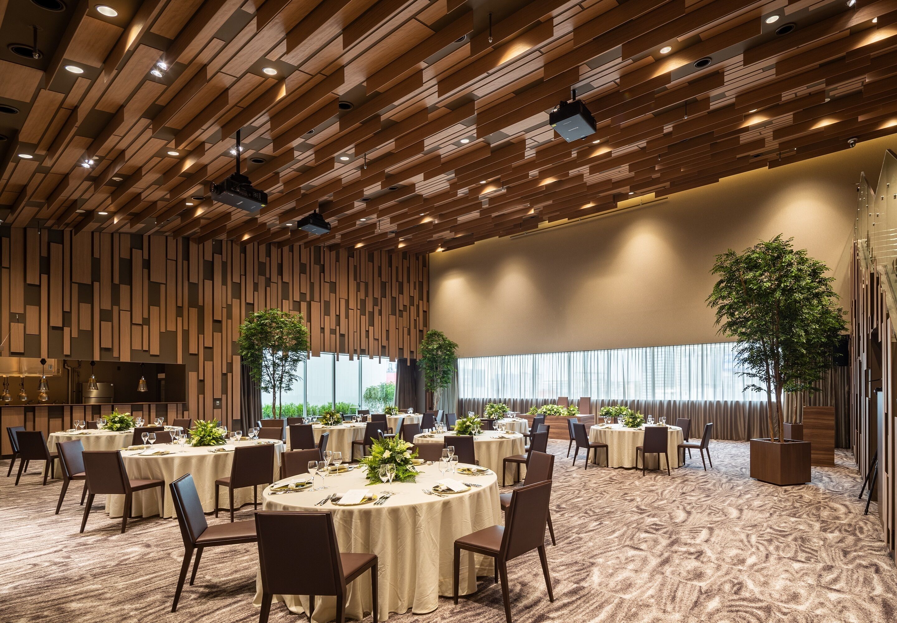 A spacious banquet hall with round tables set for dining, a high ceiling with wooden paneling, and large windows allowing natural light.