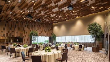 A spacious banquet hall with round tables set for dining, a high ceiling with wooden paneling, and large windows allowing natural light.