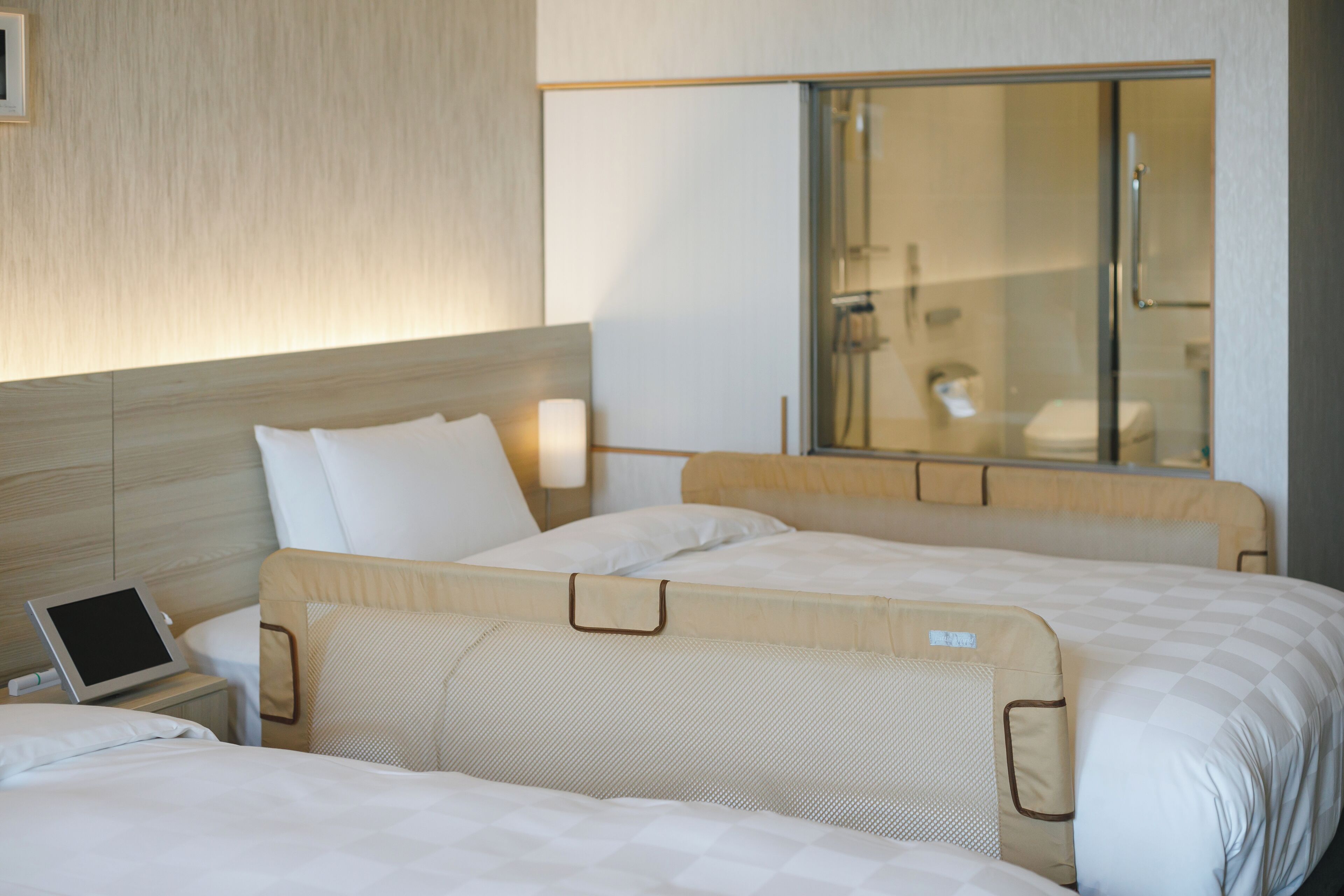 A hotel room with two beds, a bedside table with a tablet, and a large window with a view of a bathroom.