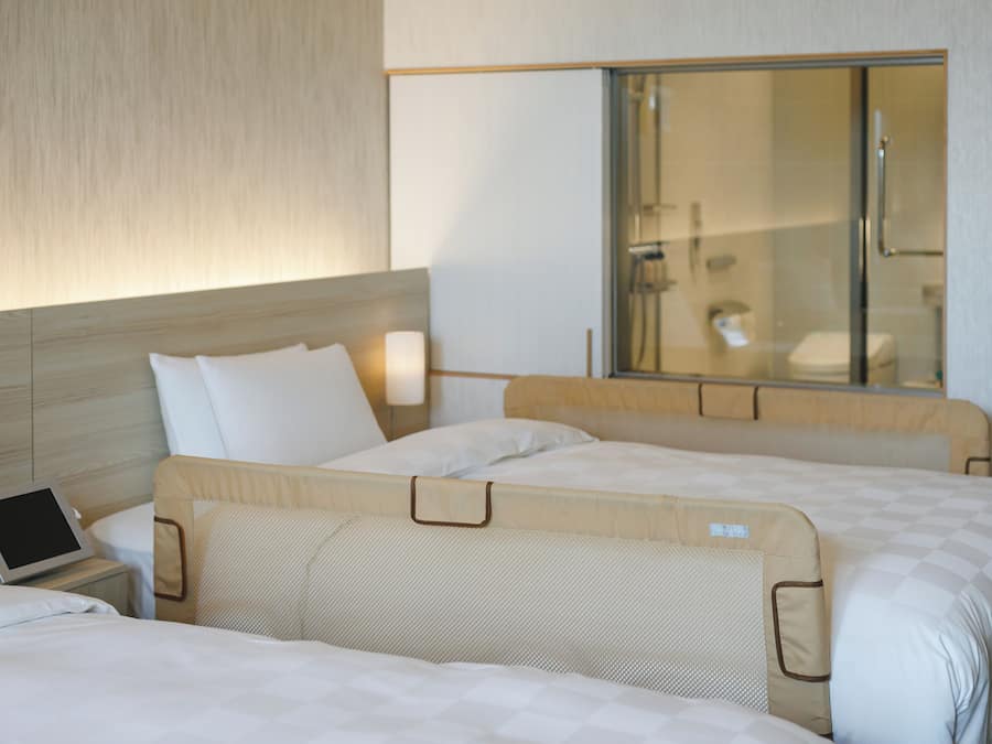 A hotel room with two beds, a bedside table with a tablet, and a large window with a view of a bathroom.
