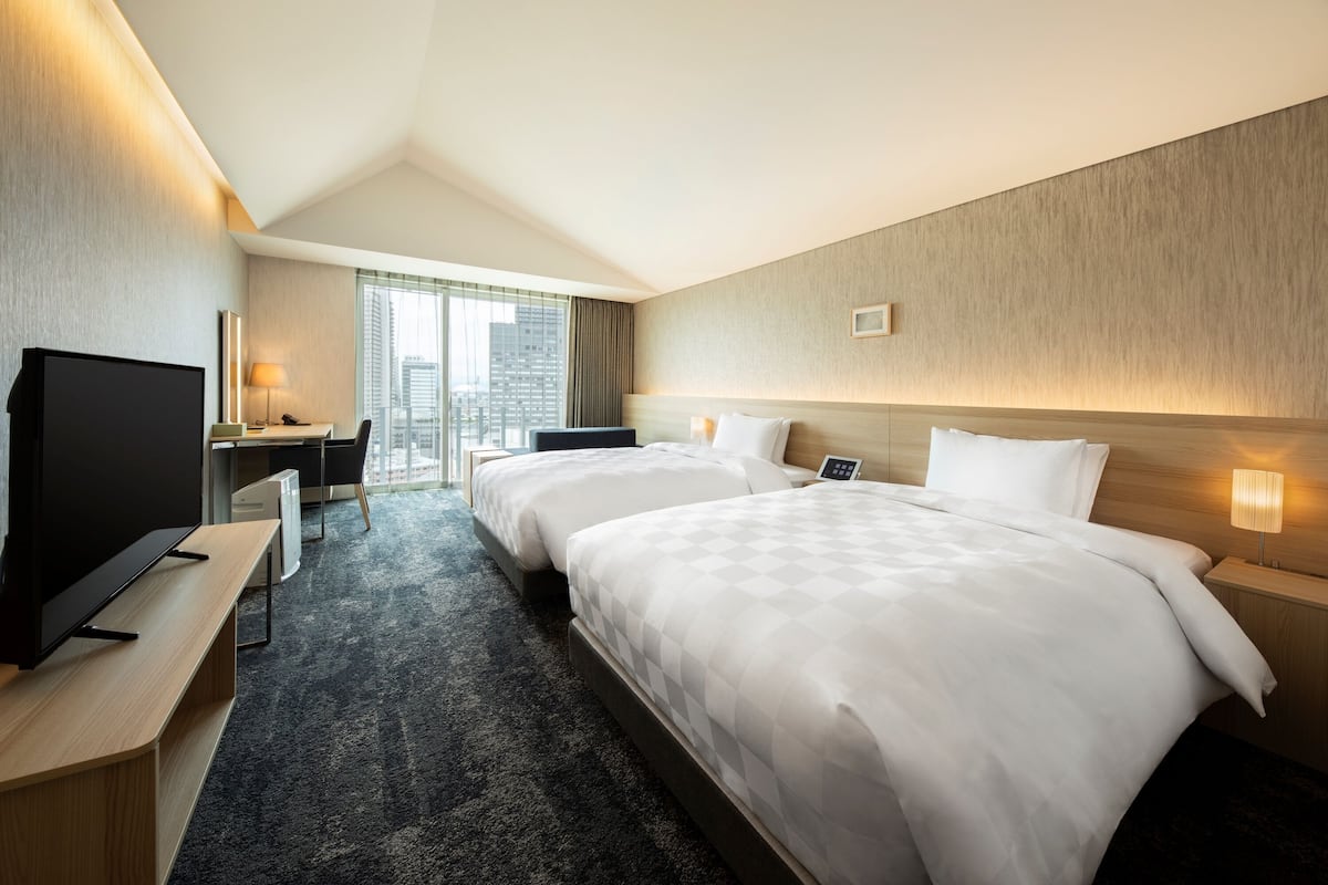 A hotel room with two beds, a desk, a TV, and a city view.