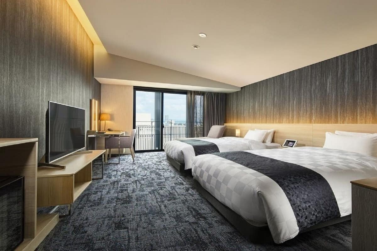 A hotel room with two beds, a TV, a desk, and a balcony with a view.