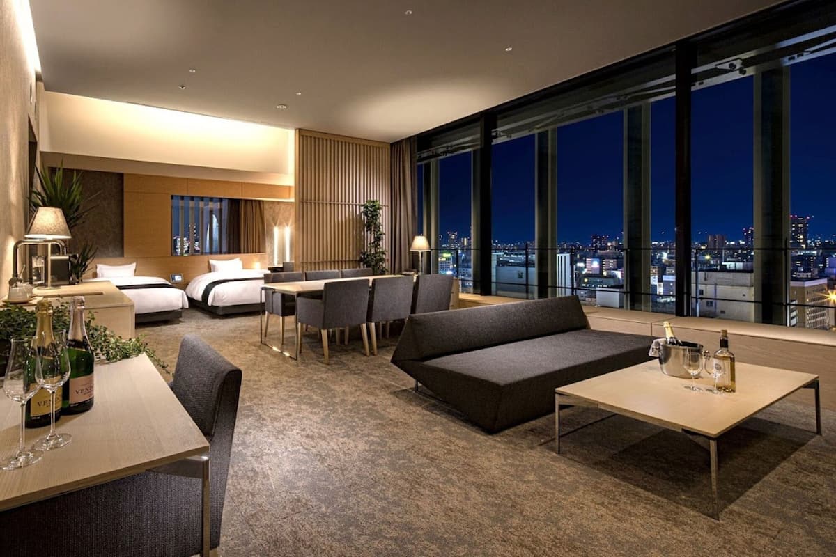 A modern hotel room with a city view, a dining area, and a seating area with a sofa and table.