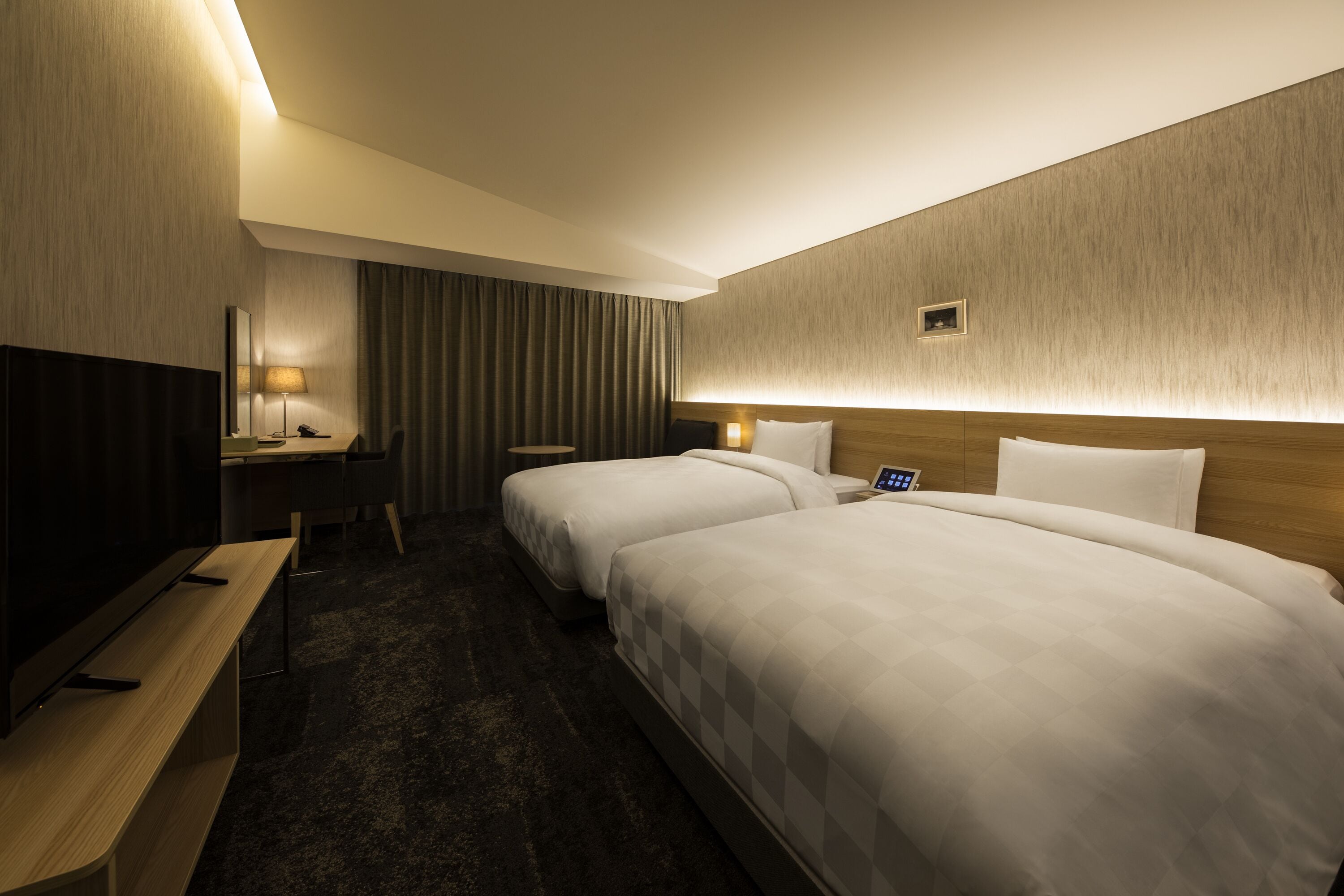 A hotel room with two beds, a desk with a chair, a TV, and a bedside table.