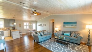 A spacious living room with a blue sofa set, a coffee table, and a patterned rug. There is a kitchen area with a dishwasher and cabinets.