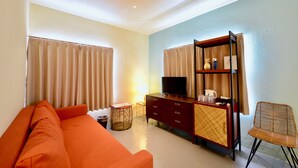A room with a sofa, a television on a wooden cabinet, a wicker chair, and a small table with a lamp. - Adan Hotel (Nago)