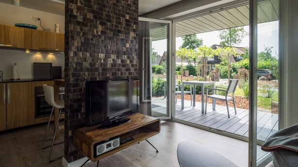 En moderne stue med et fladskærms-tv på et træstel, en stor skydedør, der fører til en udendørs terrasse, og et køkkenområde med træskabe og et indbygget ovn.