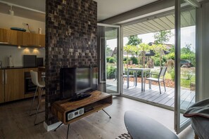 En moderne stue med et fladskærms-tv på et træstel, en stor skydedør, der fører til en udendørs terrasse, og et køkkenområde med træskabe og et indbygget ovn.