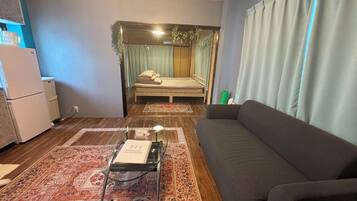 A compact living space with a sofa, a small table with a book, a bed with curtains, and a refrigerator.