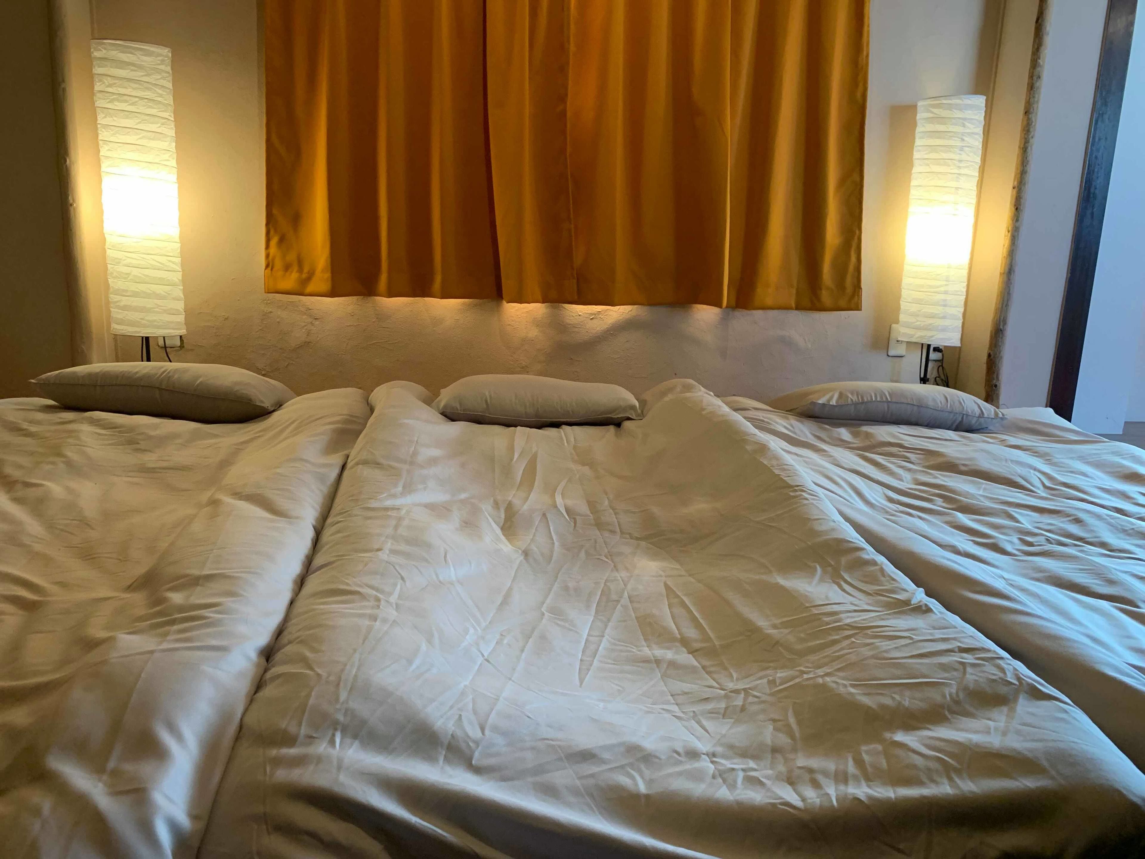 Un lit avec du linge de lit blanc et deux oreillers, une tête de lit agrémentée d’un rideau jaune, et deux lampes fixées au mur, une de chaque côté.