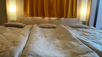 Un lit avec du linge de lit blanc et deux oreillers, une tête de lit agrémentée d’un rideau jaune, et deux lampes fixées au mur, une de chaque côté.