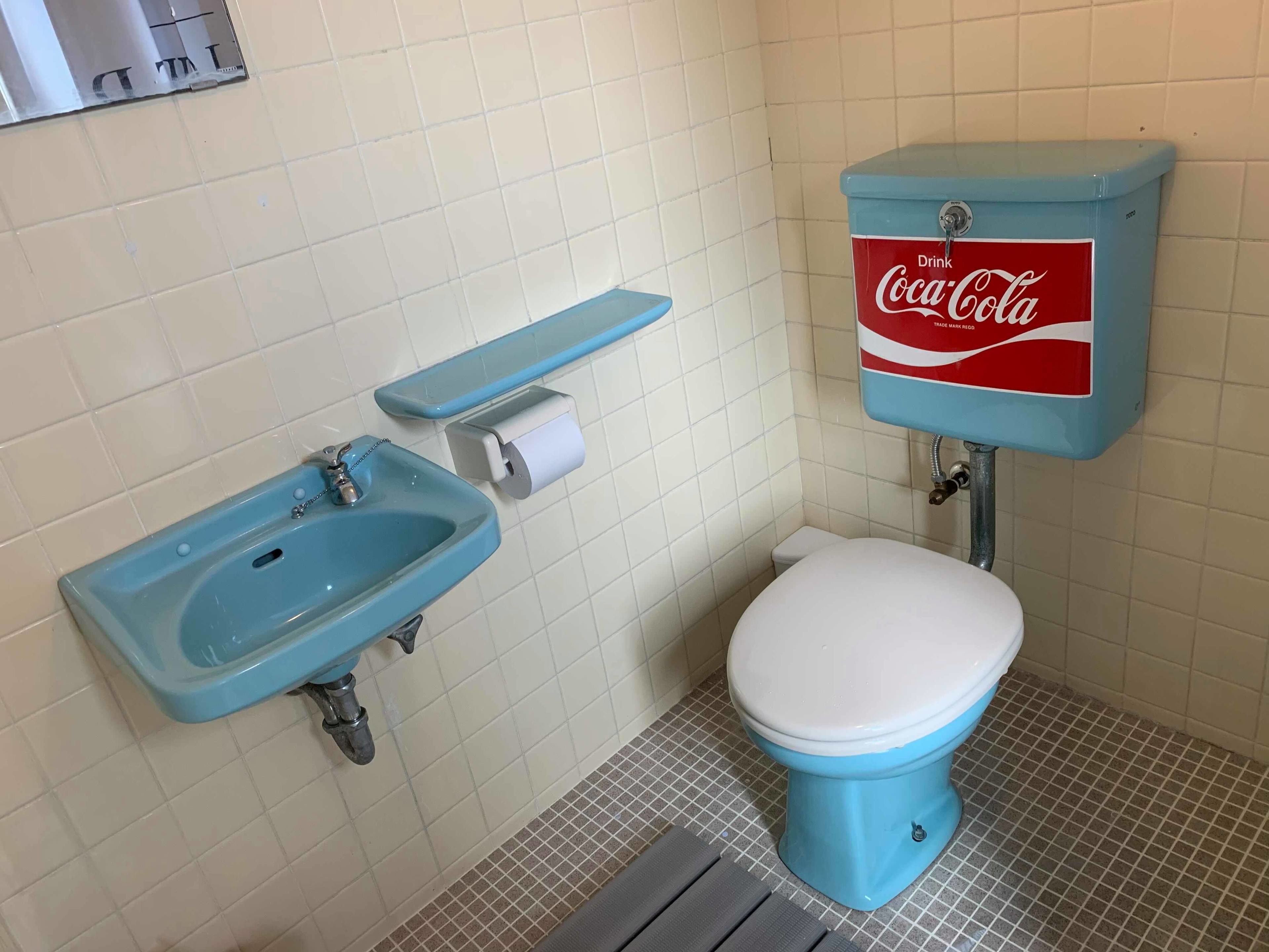 Ein Badezimmer mit blauem Waschbecken, Toilette und einem Tank mit Coca-Cola-Branding.