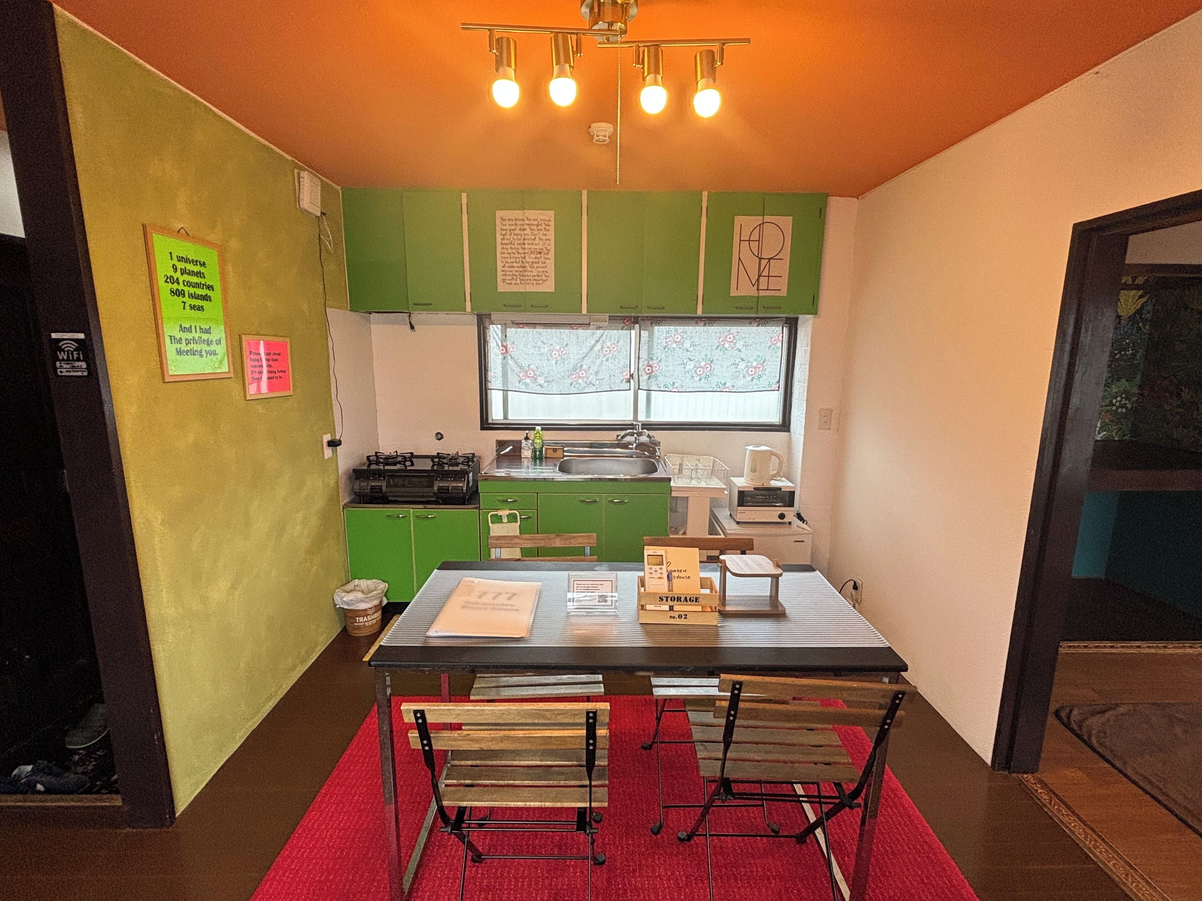 A compact kitchen with green cabinets, a gas stove, a sink, and a window with floral curtains. There is a table with chairs and a red rug.