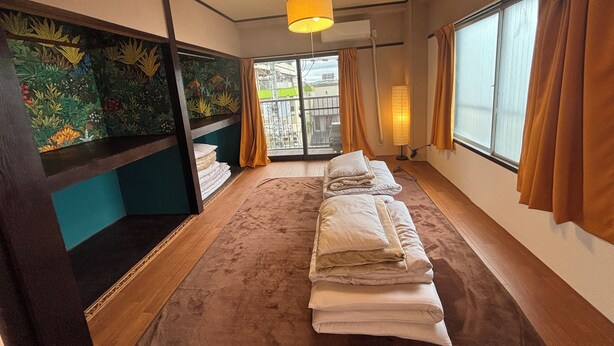 A hotel room with a bed, a window with curtains, a wall with a floral mural, and a ceiling light. - 777 Takamatsu Guest House (Takamatsu)