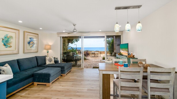 Una sala moderna con un sofá seccional oscuro, una mesa de comedor de madera con sillas, y vista a la playa a través de puertas de vidrio.