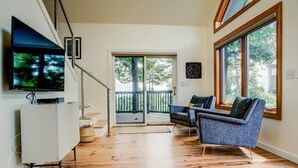 Un salón moderno con un televisor de pantalla plana, dos sillones grises, un mueble blanco y un ventanal con vistas al exterior.