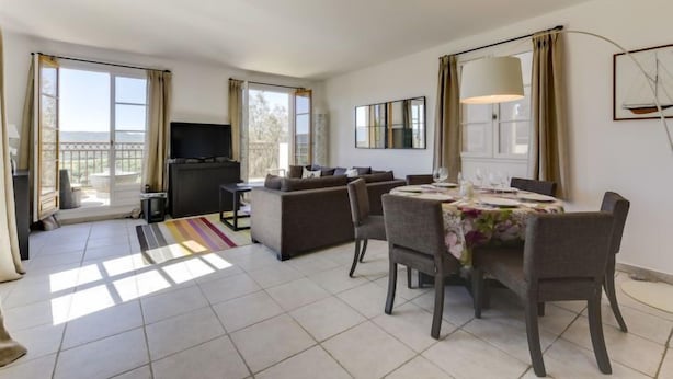 Un salón espacioso con un conjunto de sofás, una mesa de comedor con sillas, un televisor de pantalla plana y grandes ventanales con vistas al exterior.
