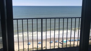 A view of the ocean through a balcony railing with beach umbrellas and a lifeguard stand visible on the beach.