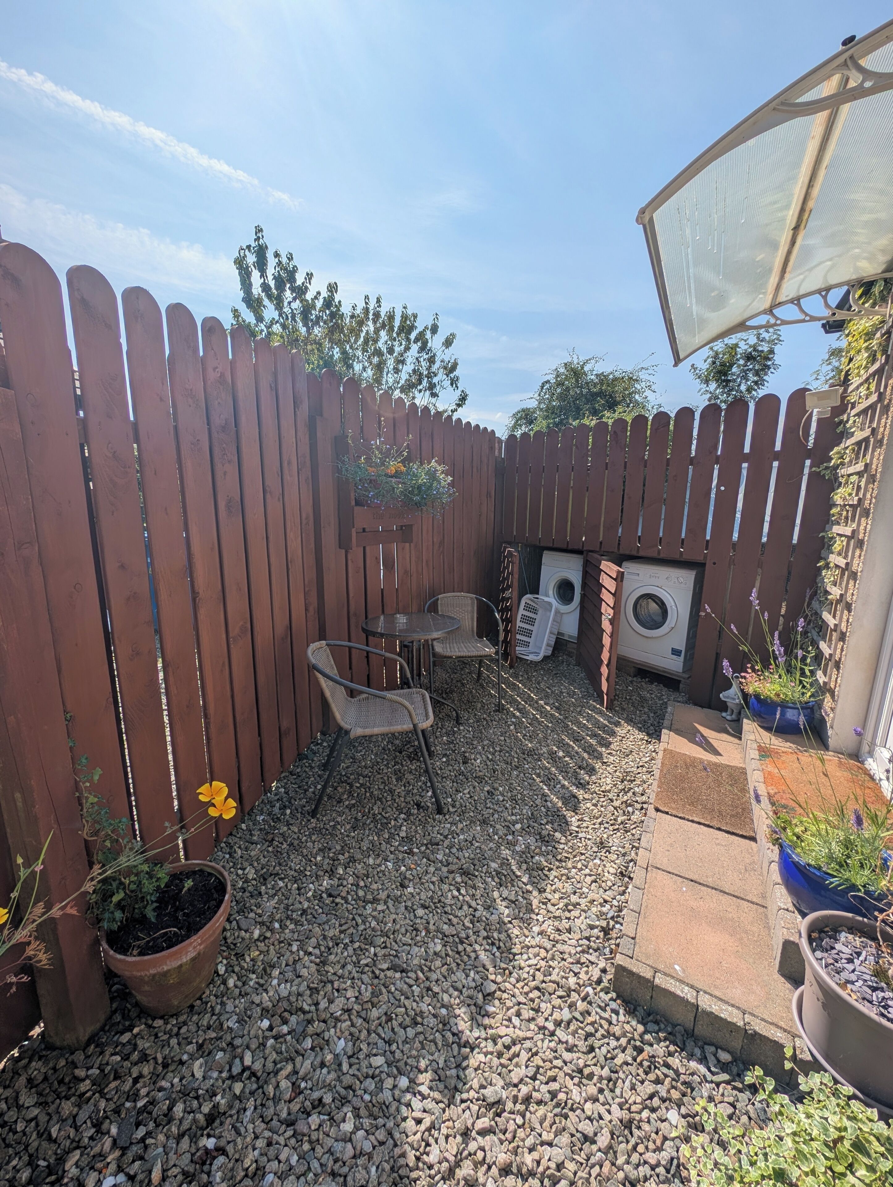 A backyard with a wooden fence, two wicker chairs, a washing machine, and potted plants.