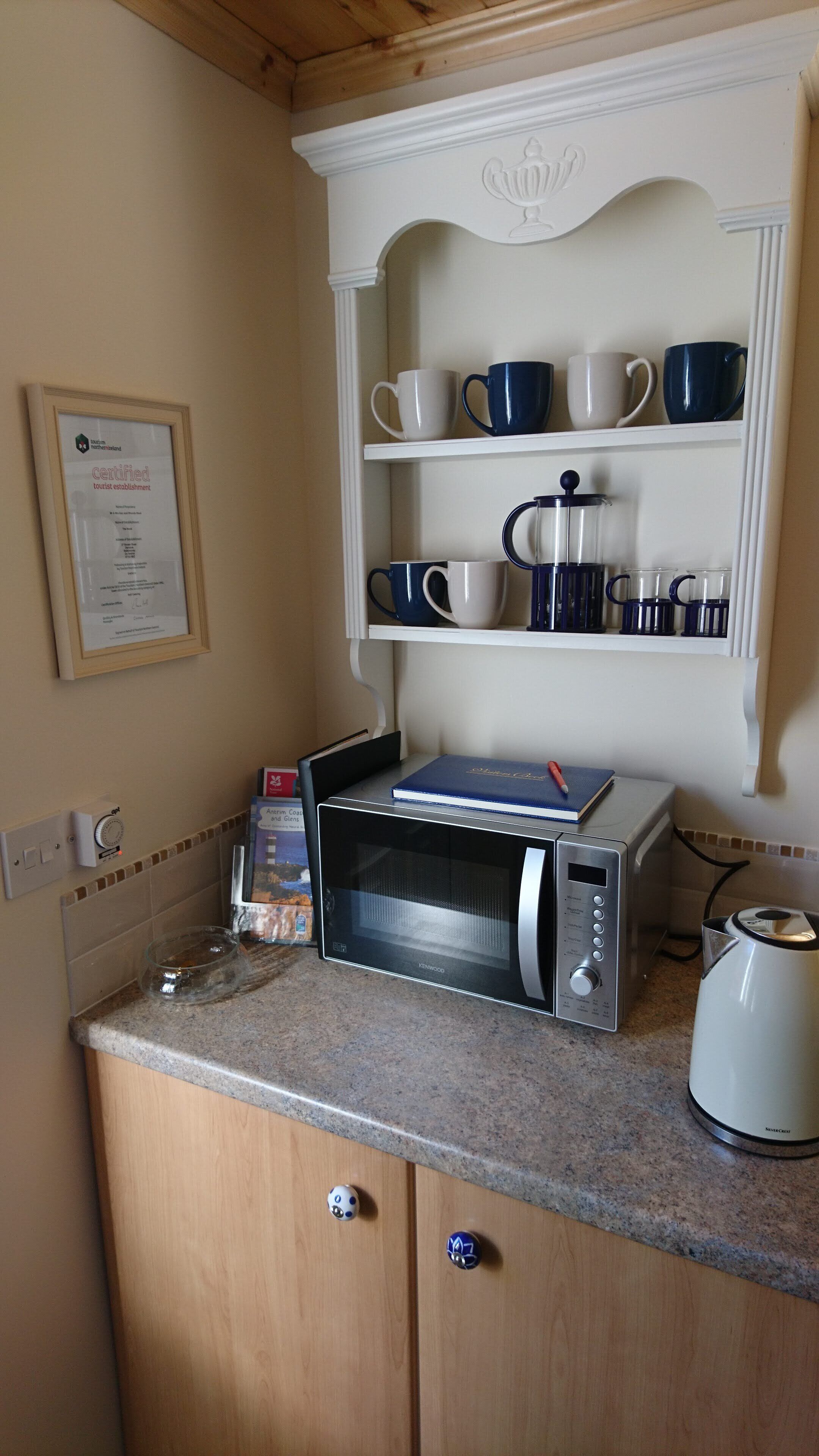 Una cocina con microondas, hervidor de agua y cafetera sobre la encimera. Se observa un certificado enmarcado en la pared y una estantería con tazas y vasos.