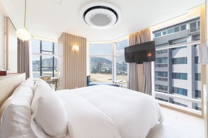 A hotel room with a large bed, a TV mounted on the wall, and a view of a cityscape through large windows. - Ocean 2 Heaven (Busan)
