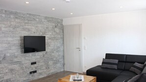 Una sala de estar moderna con un televisor de pantalla plana montado en una pared con textura, un sofá de cuero negro y una mesa de centro de madera.
