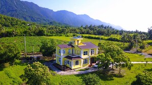 A two-story yellow house with a balcony, surrounded by lush greenery and mountains in the background. - Straybirds BnB (Donghe)