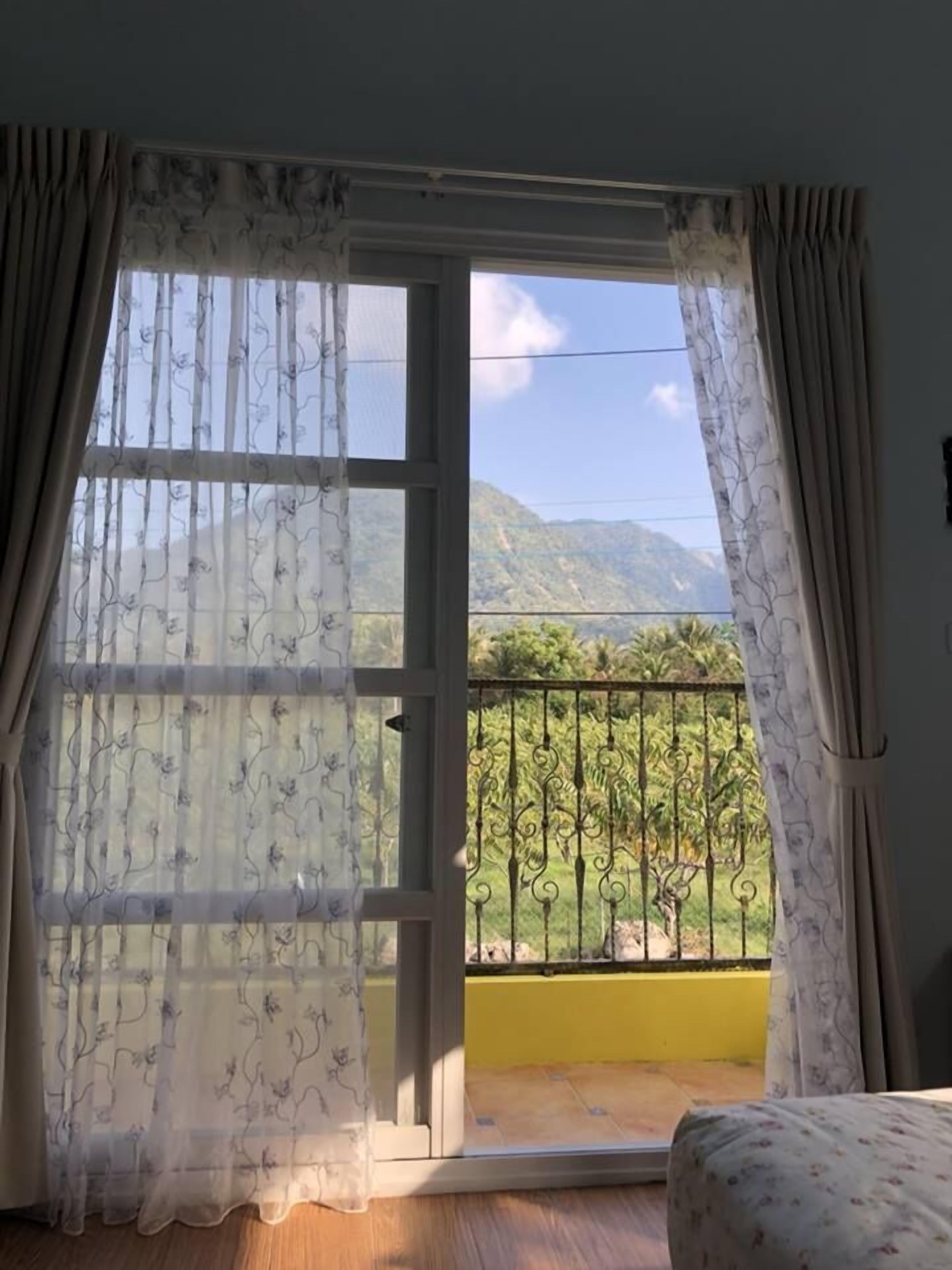 A room with a large window, curtains, and a view of greenery and mountains.