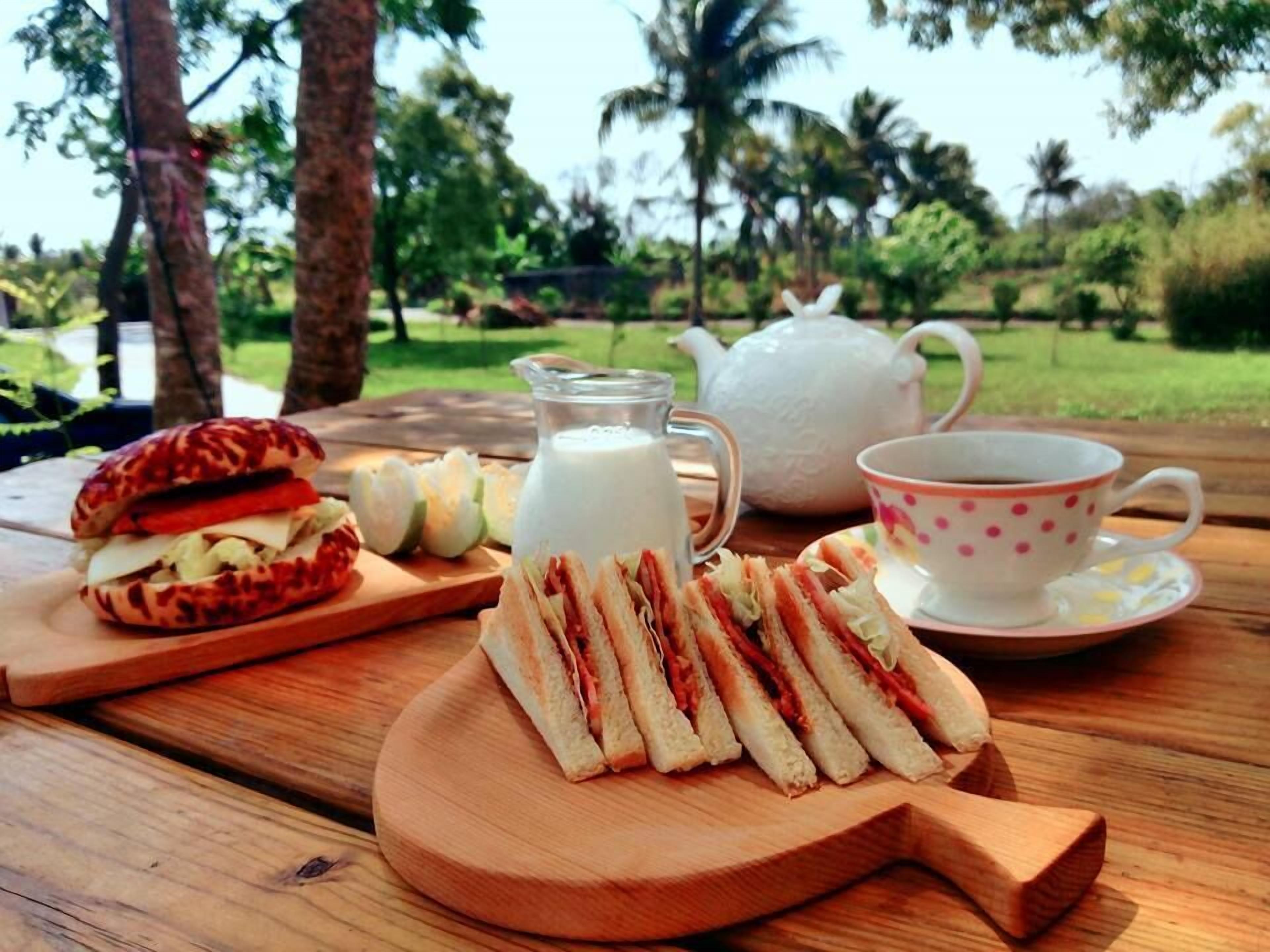 A picnic setup with sandwiches, a jug of milk, a teapot, and a cup on a wooden table in a park.