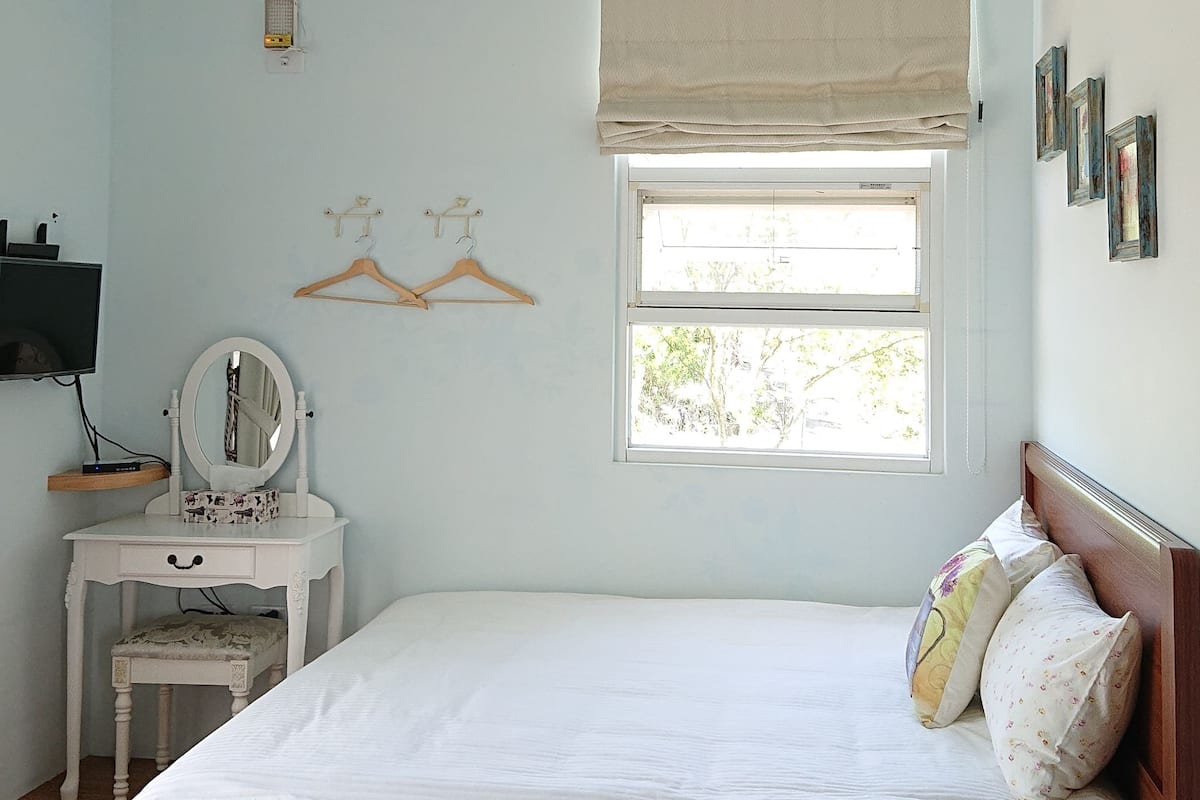 Un dormitorio con una cama de madera, una mesita de noche con espejo, una lámpara colgante, una ventana con persiana y un televisor montado en la pared.