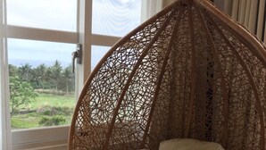 A wicker hanging chair with a cushion, positioned near a window with a view of greenery and a cloudy sky. - Straybirds BnB (Donghe)