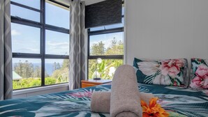 A room with a bed, a window with a view of trees and the sea, and a flower on the bed. - Bucks Point House (Norfolk Island)