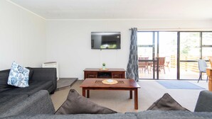 A living room with a sofa, a coffee table, a flat-screen TV, and a sliding glass door leading to an outdoor patio. - Bucks Point House (Norfolk Island)