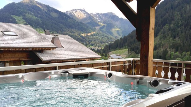 Un bain à remous avec vue sur les montagnes, une terrasse en bois et, au loin, un bâtiment de style chalet.