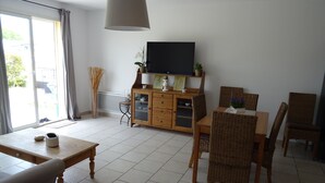 Una sala de estar con un sofá, una mesa de comedor, sillas, un televisor sobre un mueble bajo y un ventanal con cortinas.