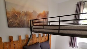 Una cama litera con cabecera de madera, una cama individual con sábana gris, una mesita de madera y un cuadro en la pared.
