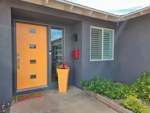 Ett modernt hus med en knallorange dörr, en liten blomkruka och ett fönster med gröna fönsterluckor.