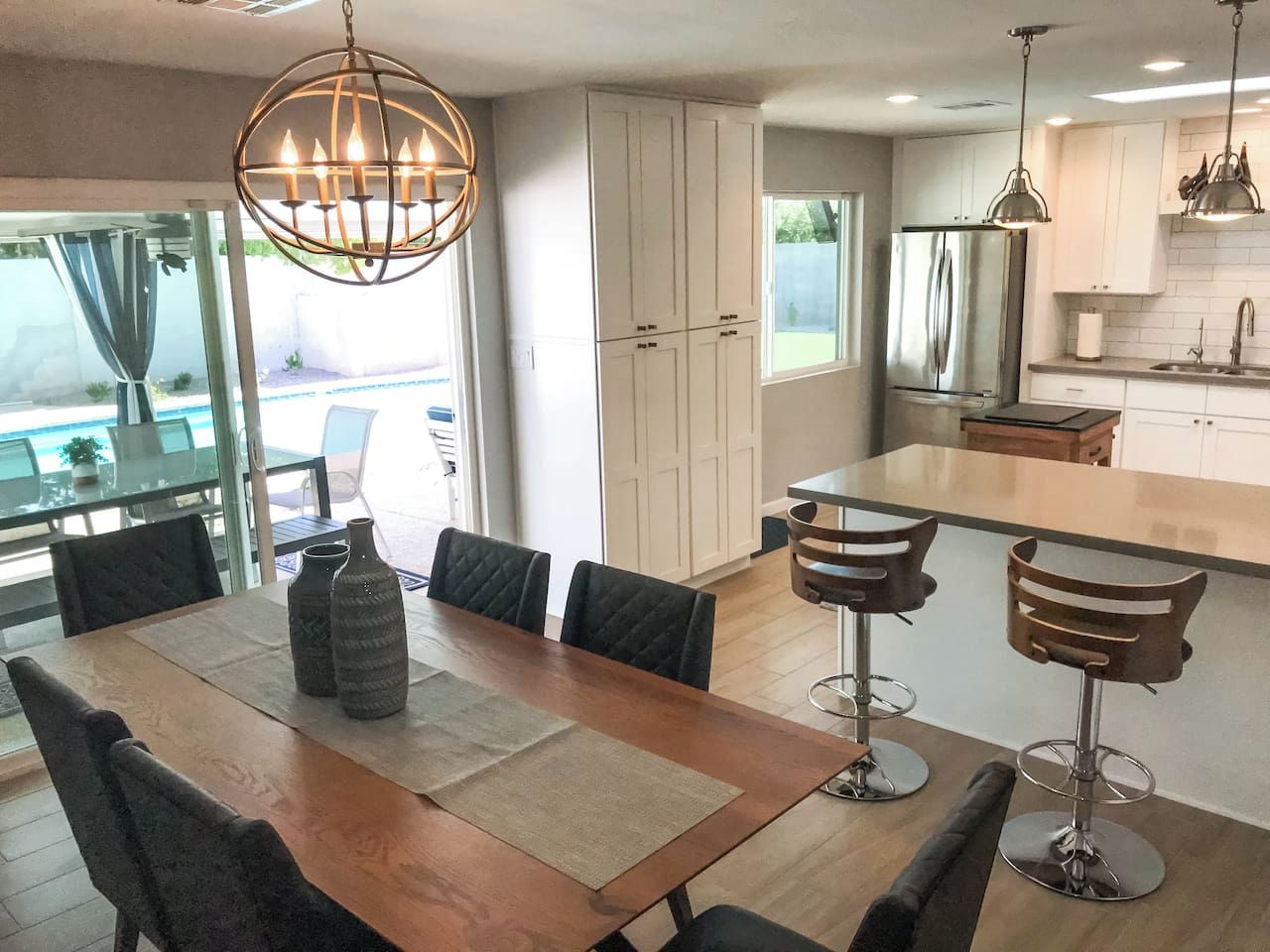 A modern kitchen with a dining area, featuring a wooden table, bar stools, and a large window.
