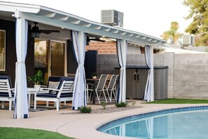 A covered outdoor seating area with chairs and a table, a pool, and a brick wall. - Pecos Lane | Poolside by Day, Wolf Den by Night (Scottsdale)