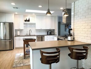 A modern kitchen with white cabinets, a stainless steel refrigerator, a central island with bar stools, and pendant lighting. - Pecos Lane | Poolside by Day, Wolf Den by Night (Scottsdale)