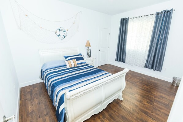 Un dormitorio con una cama grande, suelo de madera, cabecero blanco, mesita de noche con lámpara y ventana con cortinas.
