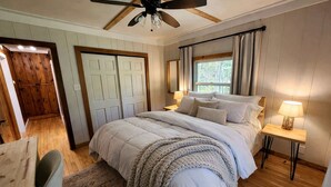 Ein Schlafzimmer mit Bett, Nachttischen, Deckenventilator, Fenster mit Vorhängen und Holztür.