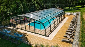 A covered swimming pool with a glass enclosure, wooden deck, and outdoor seating area.